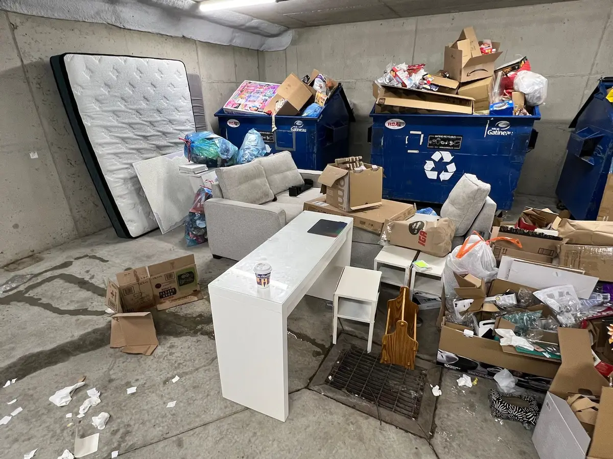 A building's garbage room with a mattress, sofa, desk and boxes left beside full recycling bins